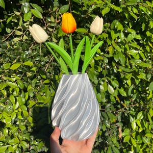 Puede incluir: Un jarrón plateado con un diseño en espiral contiene tres tulipanes artificiales. Un tulipán es amarillo y naranja, los otros dos son blancos. Las hojas verdes de los tulipanes contrastan con el jarrón plateado.