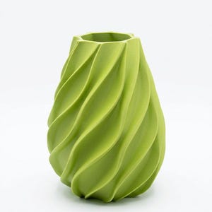 Puede incluir: Un jarrón verde con un diseño único y arremolinado. El jarrón tiene una base ancha que se estrecha hacia la parte superior, con una abertura hexagonal. El exterior presenta una serie de crestas curvas en forma de ola que envuelven el jarrón.