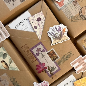 Puede incluir: Primer plano de varios paquetes de papel marrón decorados con pegatinas florales, sellos y listas de lectura. Los paquetes están adornados con pegatinas con libros abiertos, flores e ilustraciones de estilo vintage, creando un tema literario.