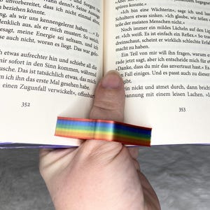 Könnte beinhalten: Ein Regenbogenfarbenes-Schleifen-Lesezeichen befindet sich zwischen den Seiten eines Buches. Das Lesezeichen besteht aus geschichteten, farbigen Bändern. Das Buch ist geöffnet, Text ist sichtbar. Eine Hand hält das Buch und das Lesezeichen.