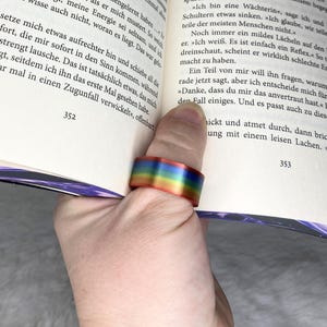 Könnte beinhalten: Ein Regenbogenfarbener Ring wird am Daumen einer Person getragen, wobei sich die Farben über die Oberfläche des Rings vermischen. Der Ring ist aus Metall und reflektiert Licht. Die Person hält ein Buch.