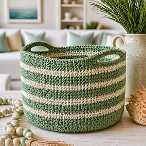 May include: A green and cream striped crochet basket with two handles. The basket is cylindrical and sits on a light-colored surface. Decorative beads and a seashell are in the foreground. The background includes a vase with greenery and a painting.