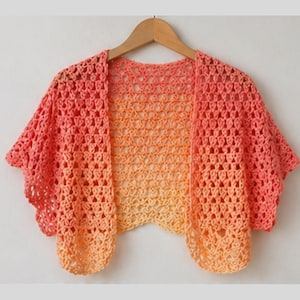 May include: A peach and orange crochet shrug with short sleeves. The open-front cardigan is made with a lacy stitch and hangs on a wooden hanger. The color gradient transitions from peach to orange to a light yellow.