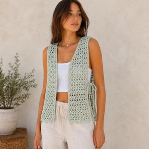 May include: A light green, open-weave crochet vest with tie closures on the sides. The vest is sleeveless and has a V-neck design. The model is wearing a white top and beige pants. The vest is a stylish layering piece.