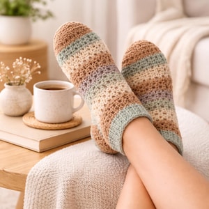 May include: A pair of handmade crochet socks in shades of brown, tan, green, and lavender. The socks are striped and have a ribbed cuff. The image shows the socks on a person's feet, with a coffee cup and a book in the background.