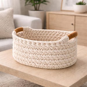 Peut inclure: Panier ovale tissé de couleur crème avec des poignées marron. Le panier est fait de fil épais et texturé et est posé sur une surface en bois clair. Les poignées sont d'un brun plus foncé, contrastant avec la couleur neutre du panier.