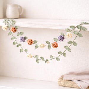 Può includere: Ghirlanda floreale all'uncinetto con un delicato design di rose e foglie. La ghirlanda presenta rose color crema, arancioni e viola, con foglie verdi. Ideale per l'arredamento della casa.