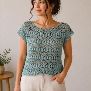 May include: A light blue crochet top with a round neckline and short sleeves. The top has a textured pattern and is worn with light-colored pants. The image shows a woman wearing the top, with a small plant and wooden table in the background.