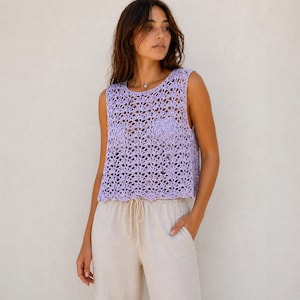 Può includere: Un top all'uncinetto lilla senza maniche con un motivo traforato. È abbinato a pantaloni beige chiaro con coulisse. L'outfit è casual e adatto alla stagione calda.