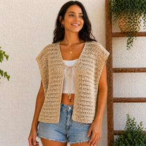 Op de afbeelding: Een beige gehaakt vest met een V-hals en striksluiting. Het open vest heeft korte mouwen en wordt gecombineerd met een witte top en een spijkerbroek. Een houten ladder en groen zijn op de achtergrond.