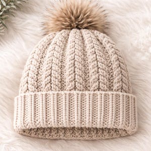 Può includere: Un berretto lavorato a maglia beige con bordo a coste e motivo intrecciato strutturato. Il cappello è sormontato da un soffice pompon in finta pelliccia marrone. Il cappello è progettato per il freddo.
