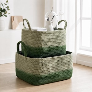 May include: Two rectangular crochet storage baskets in shades of green and beige. The baskets are stacked, with the larger one at the bottom and the smaller one on top. Each basket has two handles. The bottom portion of each basket is a darker green.