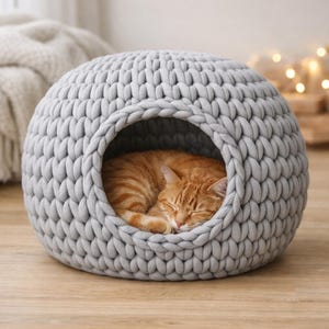 May include: A grey, knitted cat bed, spherical in shape, with a large entrance. An orange tabby cat is curled up asleep inside. The bed is made of thick, chunky yarn and rests on a wooden floor.