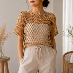 May include: A tan crochet top with a diamond pattern over a white top. The top has short sleeves and a round neckline. The model is wearing tan pants with a drawstring. The overall style is bohemian.