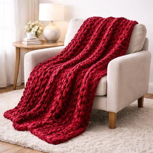 May include: A deep red knitted throw blanket, with a chunky weave, draped over a cream armchair. The blanket cascades down onto a white rug. A side table with a lamp is visible in the background, creating a cozy atmosphere.