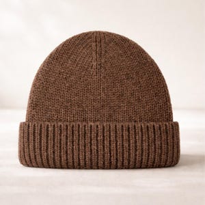 Könnte beinhalten: Eine braune Strickmütze mit geripptem Saum. Die Mütze besteht aus einem weichen, strukturierten Material und hat eine klassische, abgerundete Form. Diese warme Mütze ist perfekt für kaltes Wetter.