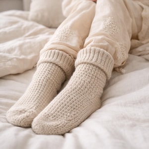 May include: A pair of cream-colored, chunky knit socks with folded-over cuffs. The socks have a textured, waffle-like pattern and are resting on a bed with cream-colored linens. The overall aesthetic is cozy and comfortable.