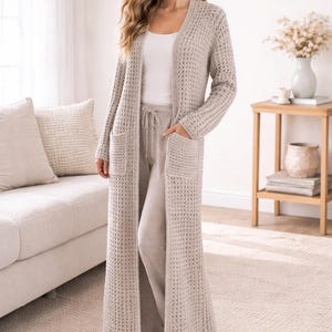 Puede incluir: Cárdigan largo de punto beige claro con dos bolsillos delanteros. El cárdigan se lleva sobre una camiseta blanca y pantalones beige claro. El conjunto se lleva en un salón con una alfombra beige y un sofá de color claro.