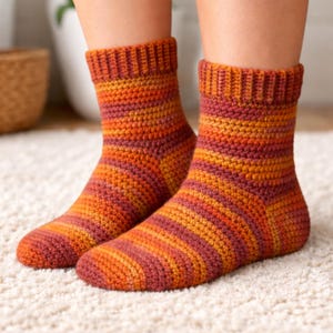 Op de afbeelding: Een paar handgemaakte, gestreepte sokken in tinten oranje, rood en geel. De sokken zijn gehaakt met een geribbelde boord en worden getoond op de voeten van een persoon, rustend op een wit, getextureerd oppervlak.