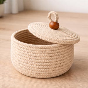 May include: A beige woven rope basket with a matching lid. The lid has a small wooden bead handle and is partially open, revealing the basket's interior. The basket is round and sits on a light-colored wooden surface.