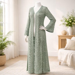 Może przedstawiać: Jasnozielony, otwarty, sięgający do ziemi kardigan zrobiony na szydełku na manekinie. Kardigan ma długie rękawy z rozkloszowanymi mankietami i dekolt w kształcie litery V. Wzór szydełkowy jest ażurowy, tworząc delikatną fakturę. Ubranie jest wystawione w dobrze oświetlonym pomieszczeniu.