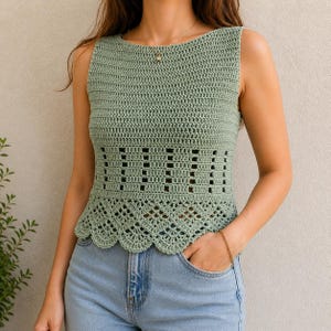 May include: A sage green, sleeveless crochet top with a round neckline. The top features a decorative openwork design across the midsection and a scalloped hemline. The handmade garment is paired with blue jeans.
