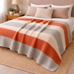 May include: A striped blanket in shades of orange, gray, and cream is draped over a bed. The blanket has a textured, ribbed design. The bed is made up with pillows and a neutral-colored headboard. A lamp and side table are visible in the background.