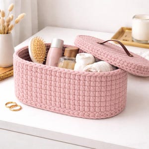 Może przedstawiać: Różowy, szydełkowy kosz do przechowywania z pasującą pokrywą. Kosz zawiera kosmetyki, w tym szczotkę do włosów, butelki i zwinięte białe ręczniki. Pokrywa ma brązowy skórzany uchwyt. Dwa złote pierścienie leżą na stole.