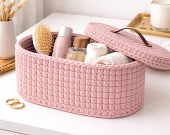 May include: A pink crocheted storage basket with a matching lid. The basket contains toiletries, including a hairbrush, bottles, and rolled white towels. The lid has a brown leather handle. Two gold rings are on the table.