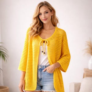 May include: A bright yellow, open-weave crochet cardigan with three-quarter sleeves. The cardigan features a tie closure at the neckline with decorative black beads. The garment is worn over a white top and paired with denim shorts.