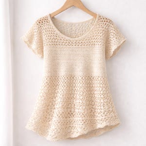 May include: A cream-coloured, short-sleeved crochet top with a scoop neck. The top features a delicate, open-weave pattern, a solid band across the midsection, and a flared hemline. It hangs on a wooden hanger.