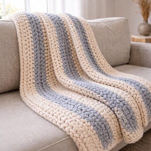 May include: A chunky knit blanket in cream and light gray stripes draped over a gray sofa. The thick yarn creates a textured, cozy appearance. The blanket's design features alternating bands of cream and light gray, offering a neutral color palette.