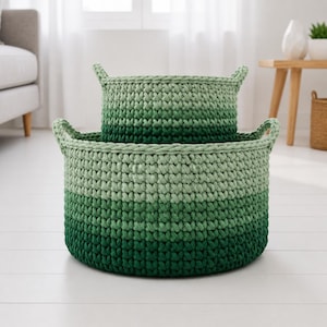 May include: Two handmade, round crochet baskets in varying shades of green. The larger basket sits beneath the smaller one, both featuring integrated handles. The baskets are made with a gradient effect, transitioning from light to dark green.