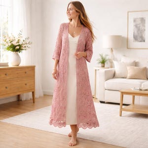 Puede incluir: Un cárdigan largo de ganchillo rosa empolvado con frente abierto y mangas tres cuartos. El cárdigan se lleva sobre un vestido blanco. El diseño de ganchillo presenta un dobladillo festoneado. El atuendo se usa en interiores.