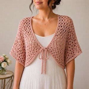 May include: A light pink crochet shrug with short sleeves and a tie closure. The openwork design of the shrug is visible. The shrug is worn over a white dress. A small bouquet of flowers sits on a table in the background.