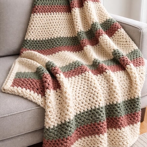 May include: A handmade, striped crochet blanket in cream, muted green, and dusty rose. The blanket is draped over a gray sofa, showcasing its textured, cozy design. The soft color palette and chunky knit suggest comfort and warmth.