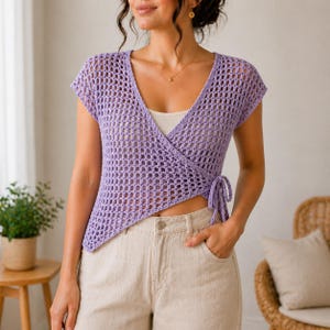 Puede incluir: Un top envolvente lila claro de manga corta con cuello en V. El top está hecho de un diseño de tejido abierto de ganchillo y tiene cierres de lazo en la cintura. La modelo lleva una camiseta sin mangas blanca debajo y pantalones cortos beiges.