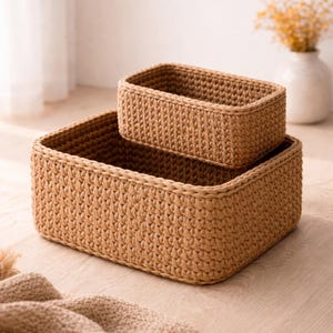 Puede incluir: Dos cestas rectangulares tejidas de diferentes tamaños, apiladas. Las cestas son de un color beige cálido y neutro, con una apariencia texturizada y de ganchillo. La cesta más grande está en la parte inferior, con la más pequeña anidada dentro. El fondo incluye un jarrón con flores.