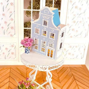May include: A miniature blue and white Dutch-style house with white trim and windows, sitting on a white table with a vase of pink flowers. The table is on a wooden floor with a herringbone pattern.