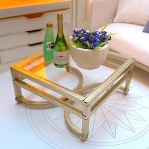 Puede incluir: Una mesa de centro de vidrio dorado con una tapa de vidrio transparente. Dos botellas, una verde y una transparente, están sobre la mesa junto con un tazón blanco lleno de flores moradas.
