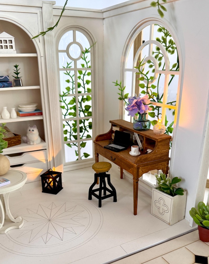 May include: Miniature dollhouse interior with a wooden desk, stool, and bookshelf. The room features arched windows with green vines, a small table, and a decorative lantern. A laptop and a cup sit on the desk. The room is painted white.