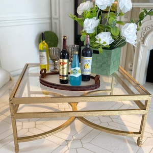 Op de afbeelding: Een gouden metalen salontafel met een glazen blad. De tafel heeft twee niveaus en een decoratief houten dienblad met drie flessen en een glas. De flessen zijn een witte wijn, een rode wijn en een blauwe frisdrank. De frisdrankfles heeft een etiket met de tekst "Limone Lago di Garda".