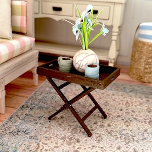 May include: A miniature dark brown wooden folding table with a tray top. The tray holds a small vase with light blue flowers, two small mugs, and a decorative vase. The table is set on a patterned rug.
