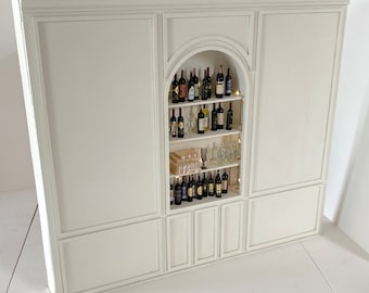 May include: A large, off-white wooden wall unit featuring a central arched alcove with three shelves. The shelves are filled with miniature wine bottles, clear glassware, and small stacked rectangular items, illuminated by integrated warm lighting.