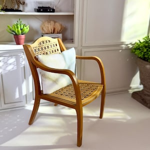 Può includere: Una poltrona in miniatura in legno con seduta e schienale in canna intrecciata. La sedia ha braccioli e gambe curve. Un piccolo cuscino quadrato con un motivo azzurro e giallo chiaro poggia sulla seduta. La sedia è in una stanza con pareti bianche.