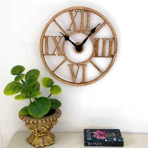May include: A brown wooden clock with Roman numeral markings and black hands. The clock is hanging on a white wall with a small potted plant and a book titled "Roses" on a shelf below.