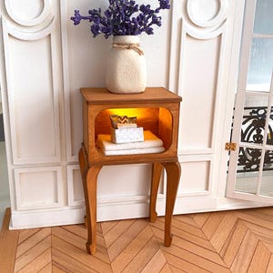 May include: A wooden side table with a shelf and three legs. The table has a light wood finish and is decorated with a vase of purple flowers. The table is sitting on a light wood floor.