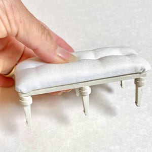 Pu&ograve; includere: Panchina bianca in miniatura con seduta trapuntata. La panca ha quattro gambe e una struttura di colore chiaro. La seduta &egrave; in morbido tessuto bianco. La panca &egrave; un modello in scala ridotta, probabilmente per una casa delle bambole o una scena in miniatura.