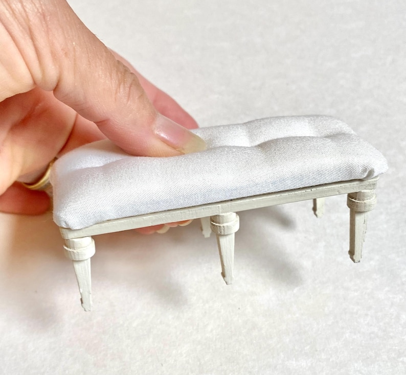 Pu&ograve; includere: Panchina bianca in miniatura con seduta trapuntata. La panca ha quattro gambe e una struttura di colore chiaro. La seduta &egrave; in morbido tessuto bianco. La panca &egrave; un modello in scala ridotta, probabilmente per una casa delle bambole o una scena in miniatura.