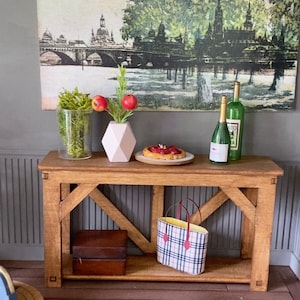 Peut inclure: Une console en bois avec un vase géométrique blanc rempli de pommes rouges, un vase en verre transparent avec des plantes vertes, une petite boîte en bois, un sac fourre-tout à carreaux et une assiette avec une tarte aux fruits. Deux bouteilles de vin sont sur la table.